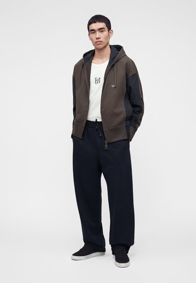 Jeune homme debout portant un sweat à capuche zippé marron foncé et noir, un t-shirt blanc avec un imprimé graphique, un pantalon noir ample et des baskets noires à semelles blanches.