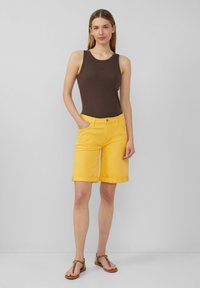 Shorts en denim jaune avec des ourlets retroussés, associés à un débardeur en côtelé marron. L'ensemble présente une coupe slim et un design classique à 5 poches.