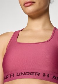 Rastezani sportski grudnjak u boji mauve s kvadratnim izrezom. Ima široku elastičnu traku s crnim natpisom "UNDER ARMOUR". Glatka tekstura.