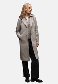 Helle graue Wollmischung Jacke mit taillierter Silhouette, ausgestattet mit einer abnehmbaren Kapuze, zwei Seitentaschen und Knopfverschluss, getragen über einer gesteppte Weste.