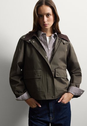 Chaqueta impermeable de color verde olivo con un cuello de pana marrón, cierre a presión y dos bolsillos frontales, superpuesta sobre una camisa a cuadros.
