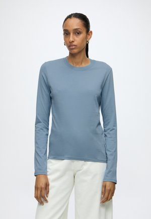 Femme debout devant un fond uni, portant une chemise bleue à manches longues et un pantalon blanc, regardant devant elle avec une expression neutre.