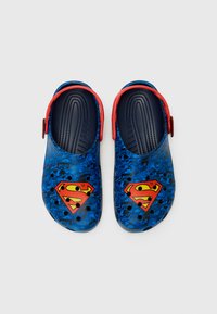 Blue rubber clogs featuring a superhero logo, red accents, and a textured footbed. The surface displays comic-style graphics with black holes for ventilation.