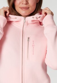 Sudadera ligera de color rosa claro con cremallera, capucha con cordón y un bolsillo frontal con cremallera. Presenta un logotipo bordado de "Sail Racing" en color a juego.