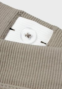 Beige ribbed fabric with a white tag featuring a transparent button. The button is accented with a brown stitch design.