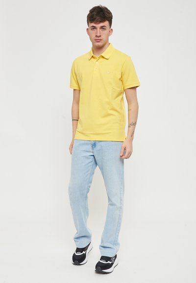 Polo giallo con una texture morbida, due bottoni e una tasca sul petto, abbinato a jeans azzurri chiari e sneakers sportive nere.