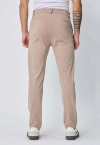 Pantalon beige à coupe droite, en tissu lisse. Dispose de deux poches arrière et d'une passante de ceinture. Associé à des chaussures blanches et noires.