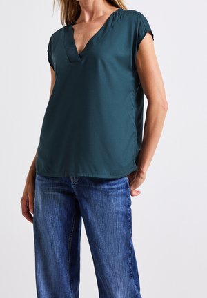 Kvinde iført en mørkegrøn V-hals bluse med korte ærmer sammen med blå denimjeans mod en ensfarvet lys baggrund.