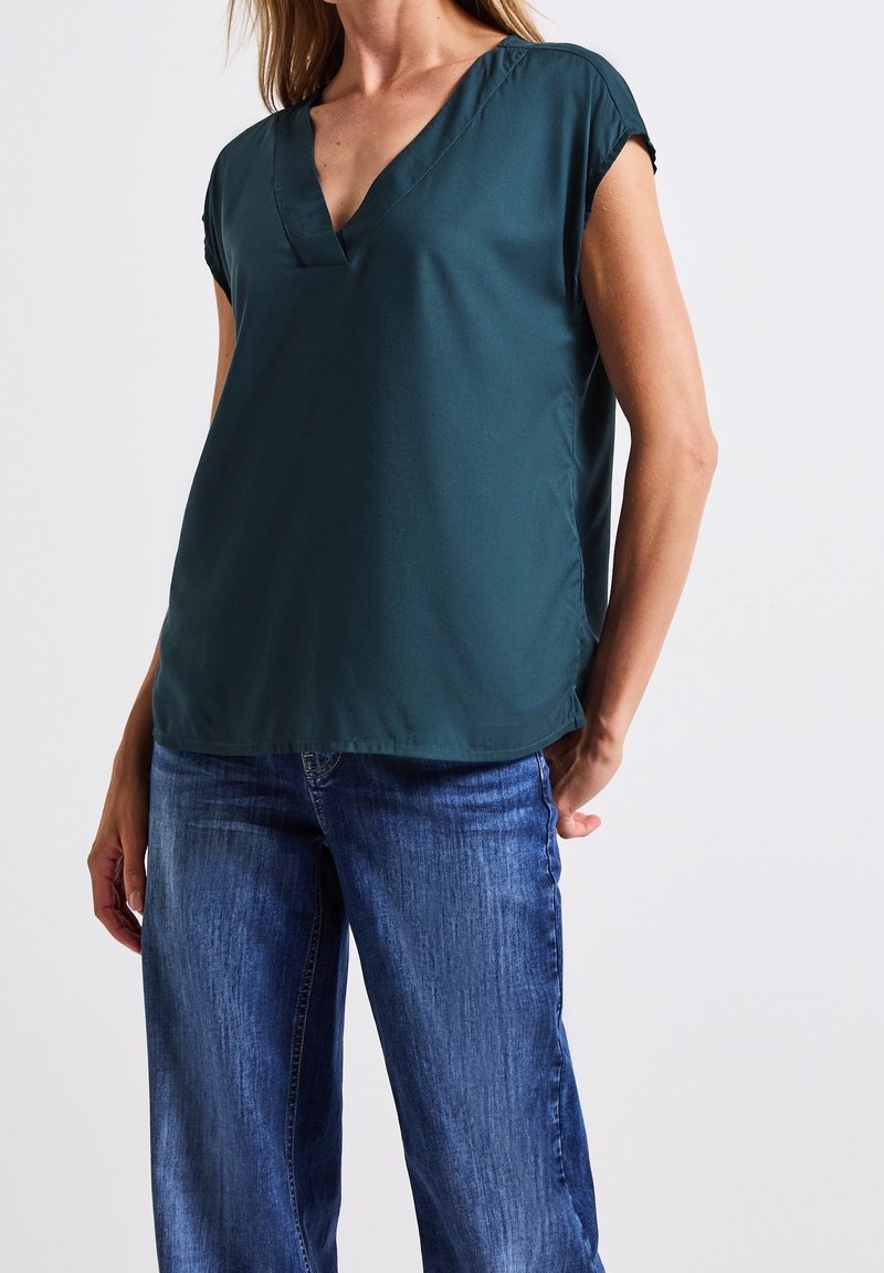 Femme portant un chemisier bleu sarcelle foncé à col en V et manches courtes, associé à un jean denim bleu sur un fond clair uni.
