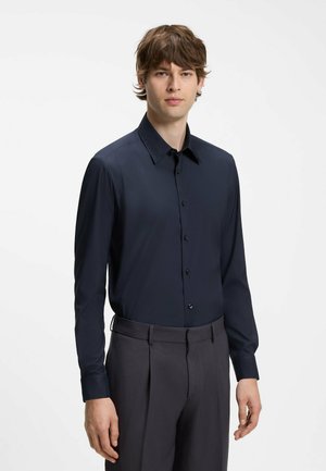 Giovane uomo con capelli castani di media lunghezza in piedi rivolto in avanti, indossa una camicia blu scuro abbottonata e pantaloni scuri su uno sfondo semplice.