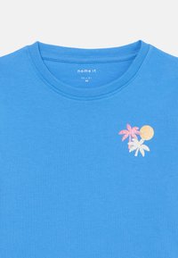 T-shirt bleu à manches courtes en coton. Présente un imprimé pailleté d'un palmier et du soleil en rose, blanc et jaune sur la zone gauche de la poitrine.