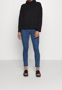 Svart stickad polotröja med ribbat nederkant, kombinerad med figurnära blå denimjeans och svarta öppna sandaler på en neutral bakgrund.