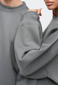 Gray sweatshirts with a ribbed neckline and cuffs, featuring a small embroidered logo. Soft texture and relaxed fit, showcasing layered sleeves.