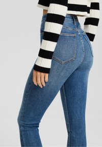 Mujer vistiendo jeans skinny de tiro alto azules y un suéter de rayas blancas y negras de manga larga y cortado, con la mano apoyada en el muslo.