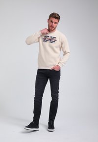 Felpa beige chiaro con un motivo grafico testurizzato blu navy sul davanti, indossata con jeans scuri e sneakers nere, in piedi di fronte a uno sfondo semplice.