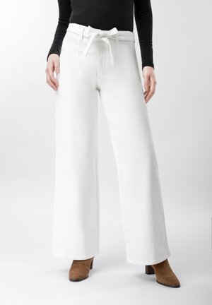 Person trägt hoch taillierte, weit geschnittene weiße Hose mit gebundenem Gürtel, schwarzen Langarmshirt und braune Stiefeletten mit Absatz.