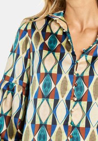 Blusa colorida con estampado que presenta un diseño geométrico en tonos verdes, azules y marrones. Hecha de tela, con cierres de botones y un cuello.