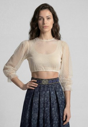 Frau mit langen welligen Haaren, die eine transparente beige kurze Bluse mit Puffärmeln und einen hoch taillierten dunkelblauen Blumenrock mit dekorativem Gürtel trägt.