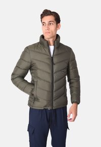 Giacca a vento verde oliva con zip frontale, colletto alto, design a rombi trapuntato, polsini elastici e tasche laterali. Indossata sopra una camicia bianca.