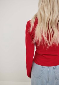 Rotes langärmliges Oberteil mit gerippter Textur, kombiniert mit hellblauen hoch geschnittenen Jeans aus Denim mit Gesäßtaschen. Haare, die den Rücken hinunterfallen.