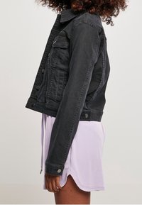 Černá džínová bunda s krátkým střihem, předními kapsami, knoflíkovými detaily a texturovaným povrchem, doplněná o světle fialový oděv pod ní.