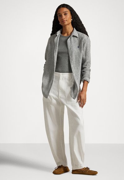 Kvinne med langt krøllete hår iført grå topp, stripete åpen skjorte, hvite høytlivsbukser og brune mokasinsko som står foran en hvit bakgrunn.