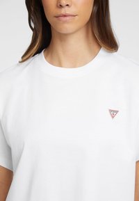 Weißes T-Shirt aus Baumwolle mit einem roten, dreieckigen "GUESS"-Logo auf der linken Brustseite. Klassischer Rundhalsausschnitt und kurze Ärmel. Glatte Textur.