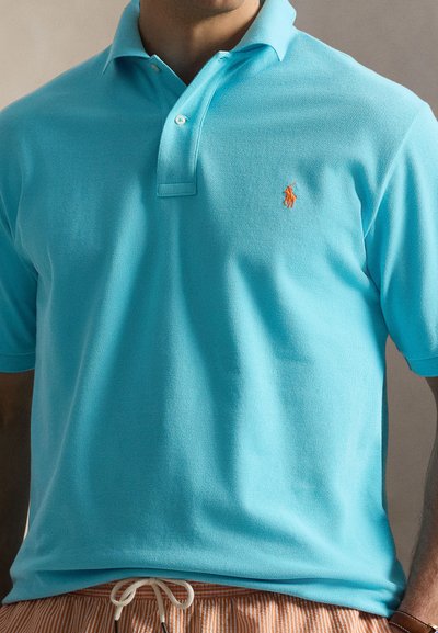 Άνδρας φοράει ανοιχτό μπλε πόλο με μικρό πορτοκαλί κεντημένο λογότυπο και ριγωτό παντελόνι με κορδόνι.