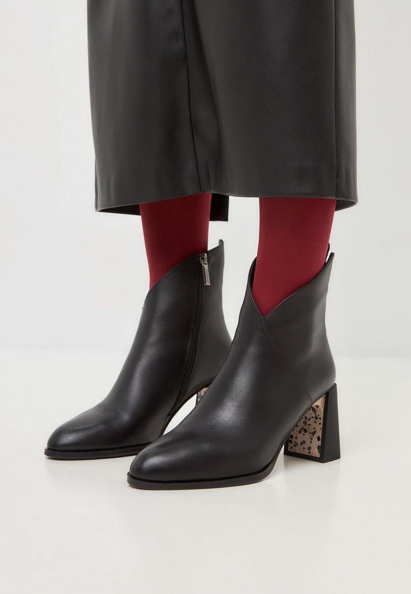 Bottines en cuir noir à talons carrés avec un motif tacheté, portées avec des collants rouges et une jupe en cuir noire.