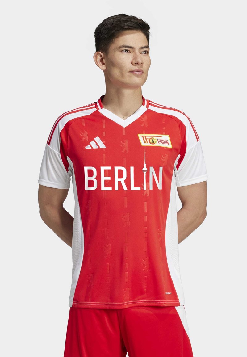 Maillot à manches courtes rouge et blanc avec un col en V, affichant "BERLIN" en lettres blanches en gras et un logo. Le motif texturé comprend des motifs d'ours.