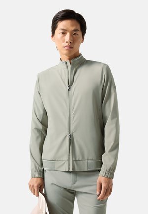 Jonge man met lichtgrijze jas met rits en bijpassende broek, houdend een lichtroze pet tegen een effen witte achtergrond.