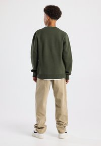 Maglione lavorato a maglia verde scuro, dal taglio rilassato, col girocollo. Abbinato a pantaloni in velluto a coste beige e sneakers bianche, visto da dietro.