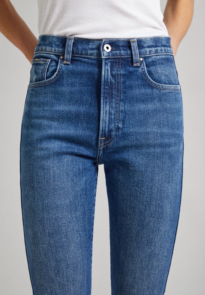 Jeans de mezclilla azul de cintura alta con un diseño de cinco bolsillos, ligero desgaste y costuras de cobre. Textura suave, ajuste slim.