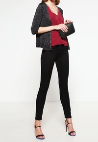 Svarta skinny jeans kombinerat med en djup burgundy top och en mörk, strukturerad cardigan. Svart kuvertväska och sandaletter med remmar och blå accenter.