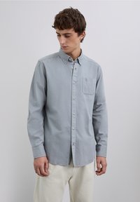 Lysegrå skjorte med knapper, lange ærmer, enkelt brystlomme, blød tekstur og afslappet pasform, kombineret med lyse bukser.