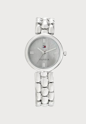 Reloj de pulsera plateado con una correa de metal enlazada, esfera redonda, marcadores de horas mínimos y el logo de Tommy Hilfiger sobre un fondo gris claro.