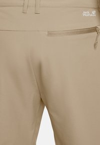 Pantalon beige Jack Wolfskin avec une poche zippée sur le côté droit et un logo de la marque visible près de la ceinture.