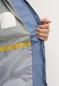 Blauwe jas interieur met gaasvoering, grijze sectie, gele accentband en een label. Heeft drukknopen aan de rand.