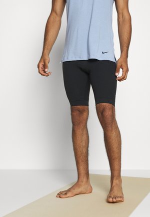 Man standing barefoot on light mat wearing light blue sleeveless top and black fitted shorts against white background.