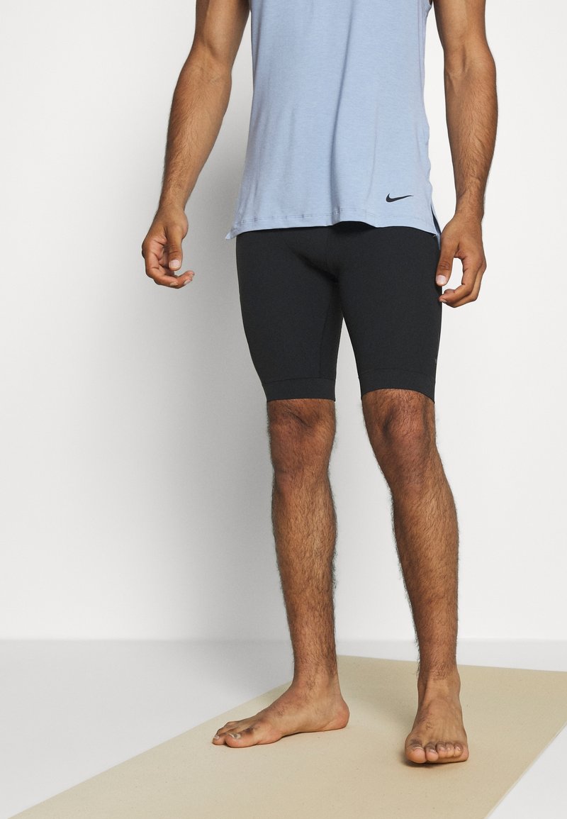 Homme debout pieds nus sur un tapis clair, portant un haut sans manches bleu clair et un short noir ajusté, sur fond blanc.