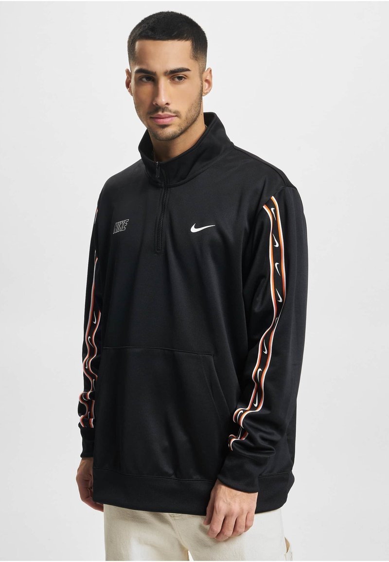 Mladý muž s krátkymi tmavými vlasmi, oblečený v čiernej mikine Nike so zipsom do štvrťky, s logom na rukávoch a prednom vrecku, stojaci pred bielym pozadím.
