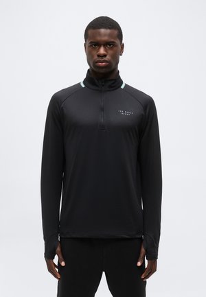 Mężczyzna noszący czarną, długorękawową sportową bluzę z zamkiem do ćwiartki i logo "Ted Baker Sport" na piersi, stojący na jednolitym tle.