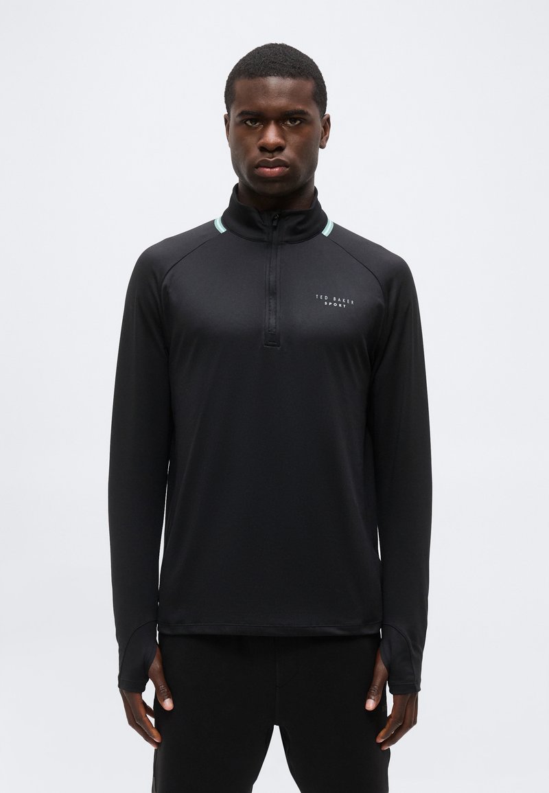Άνδρας που φοράει μακρυμάνικη αθλητική μαύρη μπλούζα με φερμουάρ μέχρι το στήθος και το λογότυπο "Ted Baker Sport" στο στήθος, στέκεται μπροστά σε ένα απλό φόντο.