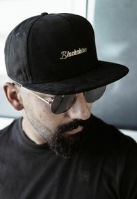 Gorra de béisbol de ante negra con "Blackskies" bordado en dorado, combinada con gafas de sol redondas plateadas y una camiseta negra.
