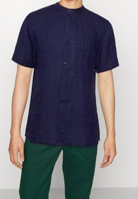 Short-sleeve, navy linen shirt with a mandarin collar, featuring front button closure and a chest pocket. Worn with green pants.
