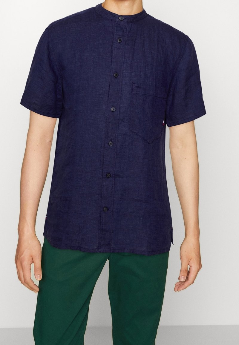 Short-sleeve, navy linen shirt with a mandarin collar, featuring front button closure and a chest pocket. Worn with green pants.