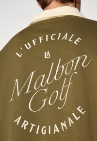 Oliven grøn polo skjorte med en kremfarget krage og brodert hvit tekst: "L’UFFICIALE LA Malbon Golf ARTIGIANALE" på baksiden.