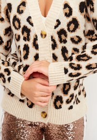 Cardigan lavorato a maglia in crema con stampa leopardata nera e marrone. Presenta un profondo scollo a V, polsini a coste e un grande bottone sul davanti.