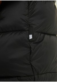 Sort puffer vest med quiltet design, glat tekstur og et lille stofmærke med en hvid firkantet accent.
