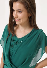 Haut vert émeraude avec un empiècement fluide et transparent. Présente un décolleté en V et est orné de perles vertes dans un motif floral sur le devant.